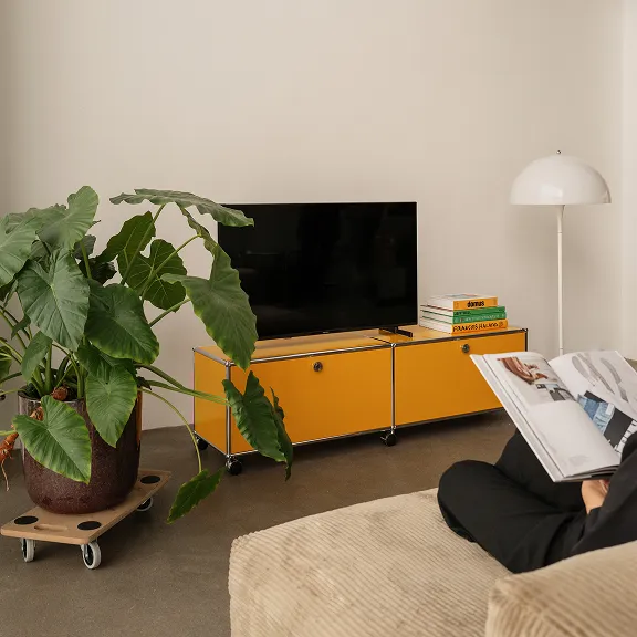Goldgelbes USM Haller Lowboard mit zwei Klapptüren und Rollen. Darauf steht ein Fernseher. 